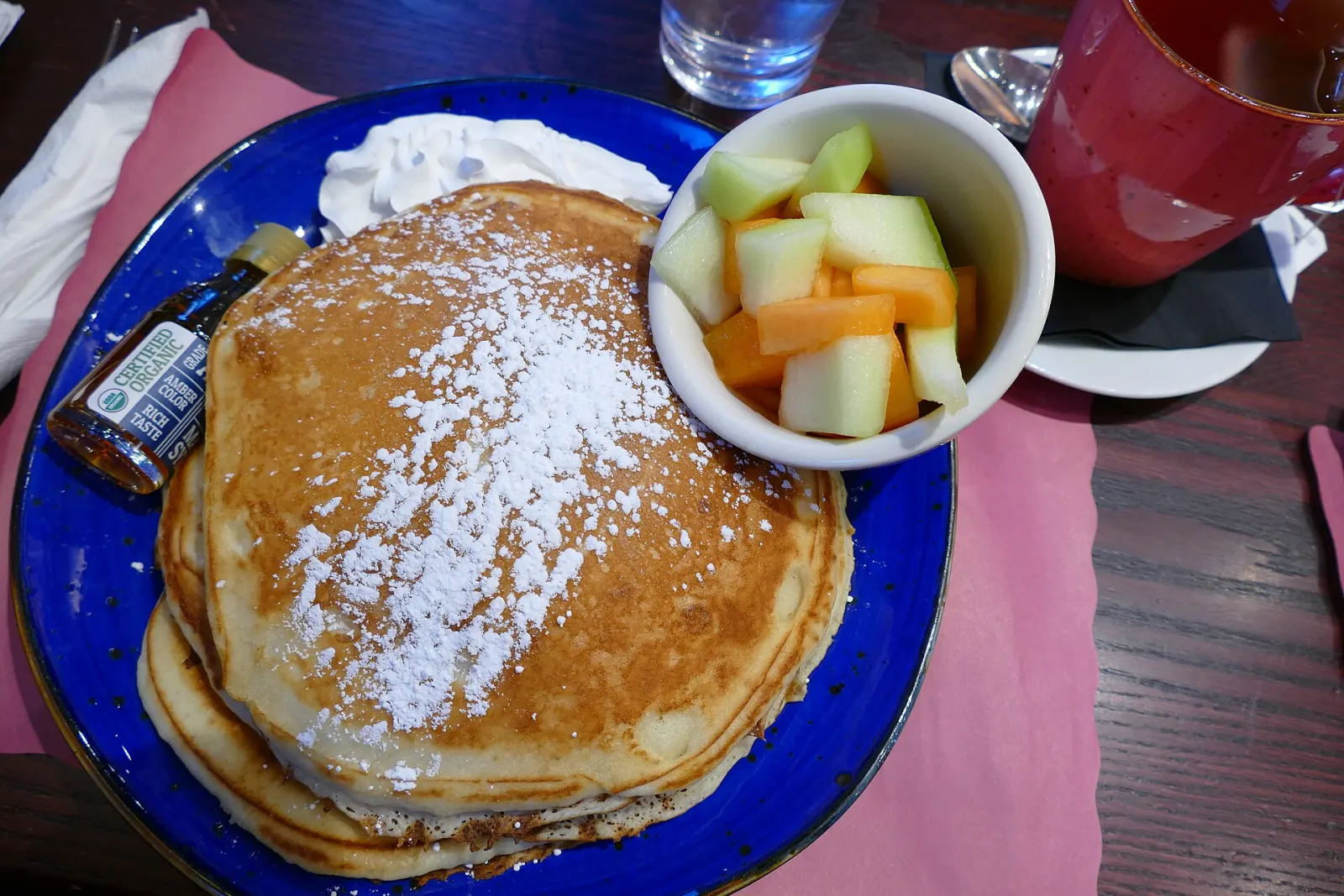 A stack of pancakes with powdered sugar, fruit and coffee at a breakfast table. The photo was not taken in Wesley Chapel.