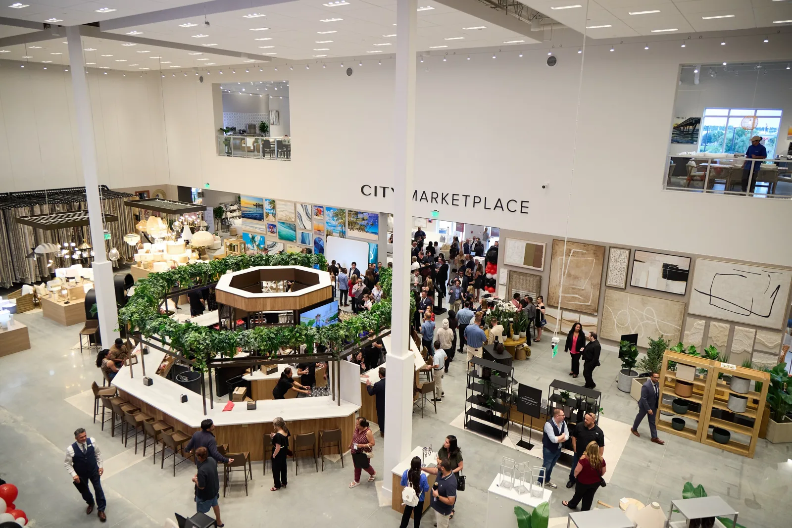 Shoppers walk through the two-story sales floor of City Home's Wesley Chapel showroom, with a cafe counter wrapped in greenery and a wall sign reading City Marketplace.